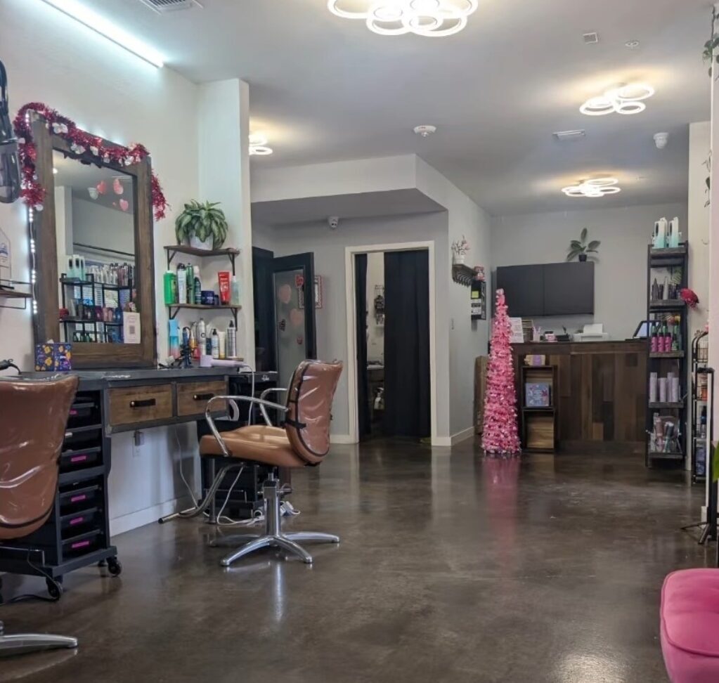 Interior of a modern hair salon: styling stations with brown chairs and mirrors on the left, product shelves, a reception area with dark wood counters on the right, and decorative pink accents including a pink Christmas tree and pink seating.