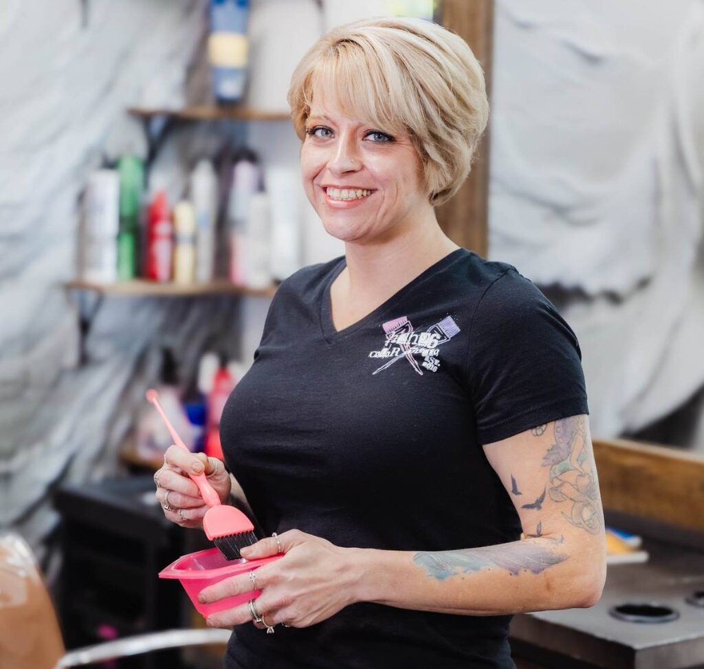 Smiling woman hairstylist in a black t-shirt mixes hair dye in a pink bowl with a brush in a salon.