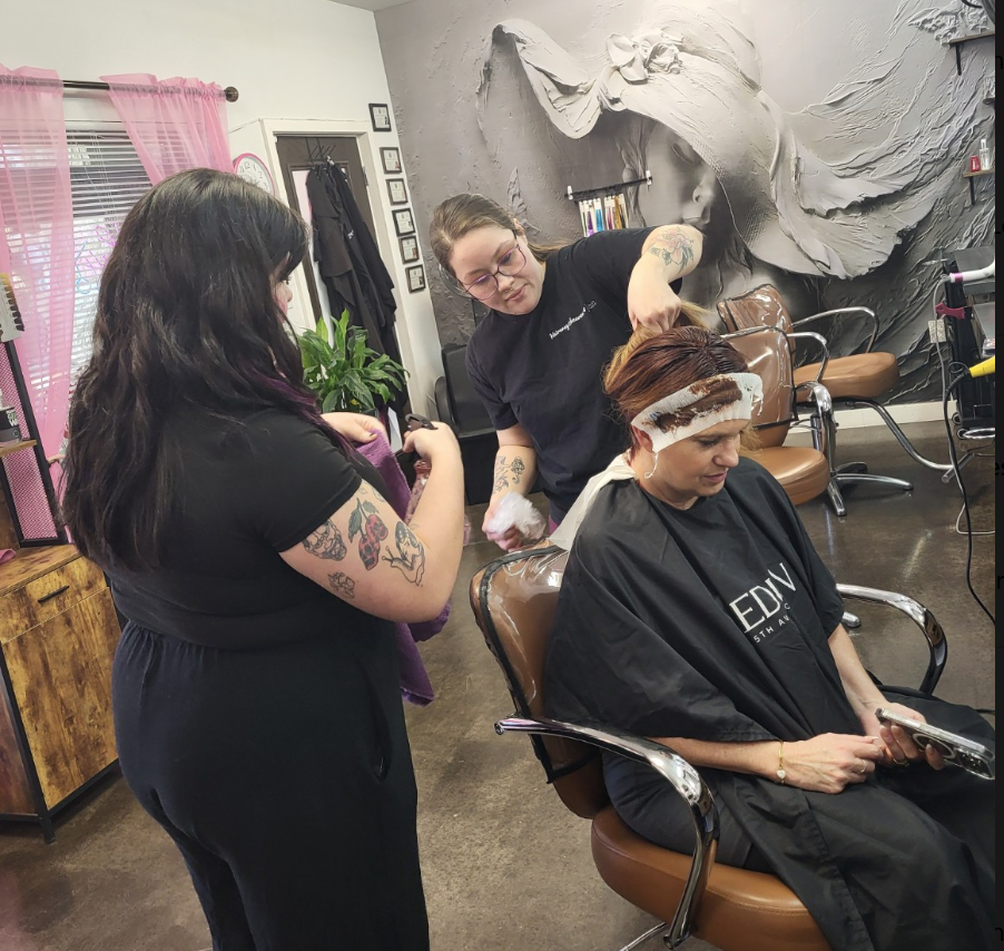 Salon scene with two stylists attending a seated client; one applies a hair treatment while the other holds a purple cloth, in front of a large mural.